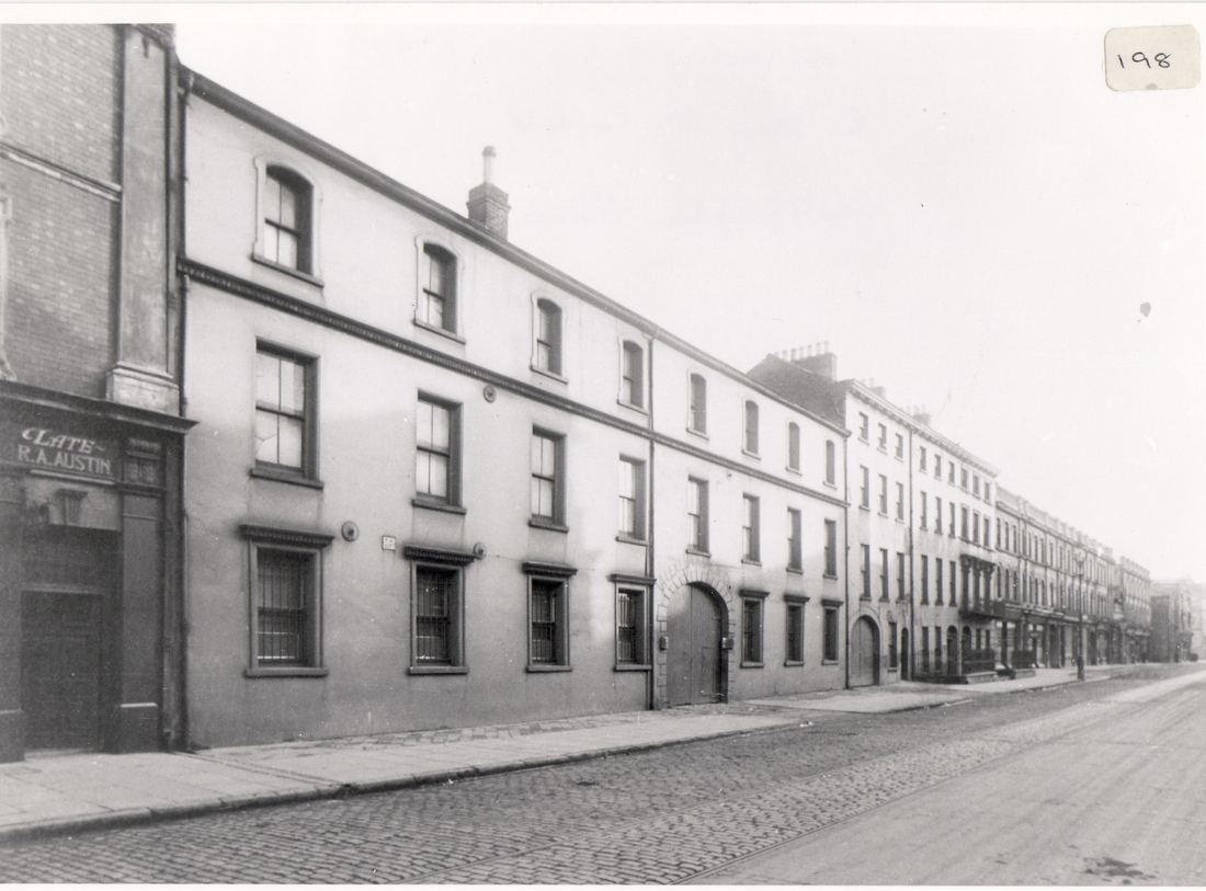 Black and white photograph of a long three-storey terrace on a cobbled street, with an arched carriage entrance